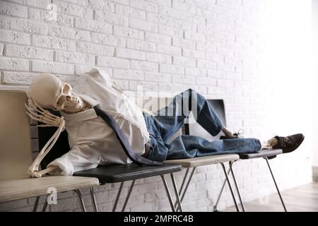 Human skeleton in office wear lying on chairs near brick wall indoors Stock Photo