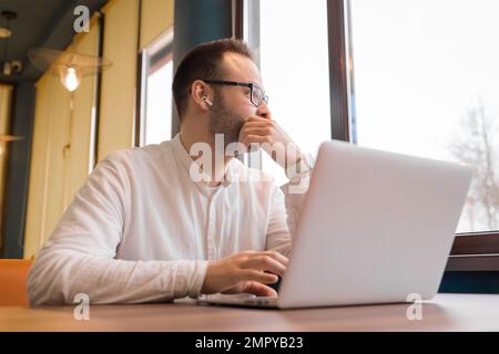 Thoughtful, stylish businessman of European appearance in glasses, with a beard, in a white shirt looking out the window distracted from work in a lap Stock Photo