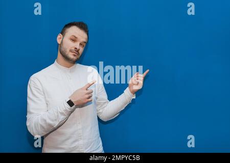 Stylish, young fellow businessman from Europe in a white shirt makes gestures or points his hands at the place for text and design on a blue backgroun Stock Photo