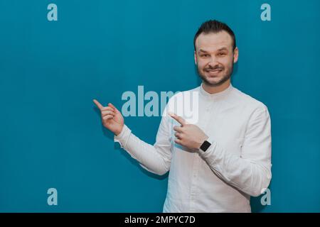 Stylish, positive fellow businessman from Europe in a white shirt makes gestures or points his hands at the place for text and design on a blue backgr Stock Photo