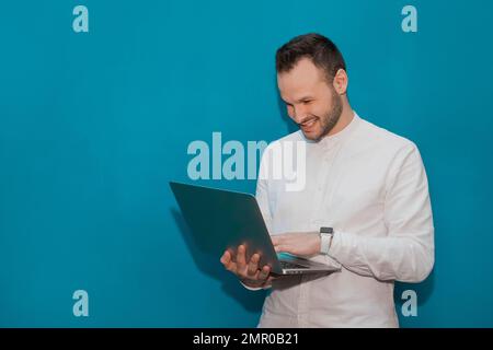 Stylish cheerful and positive businessman in a white shirt works in a laptop on a blue background. Stock Photo