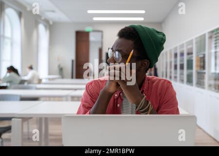 African student guy distracted from study, sitting at table with laptop in library and looking aside Stock Photo