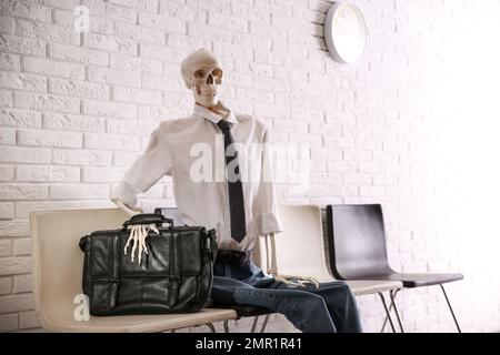 Human skeleton in office wear sitting on chair near brick wall indoors Stock Photo