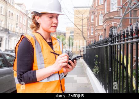 Young adult chartered civil engineer woman taking notes on her smart phone with an electronic pen, construction site, hard hat and orange personal pro Stock Photo