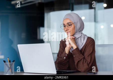 Successful smiling Arab woman in hijab working inside modern office, Muslim woman using laptop at work, business woman satisfied with achievement results typing on computer keyboard. Stock Photo