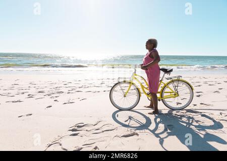 Side view of african american senior woman with bike standing on sandy beach against clear sky Stock Photo