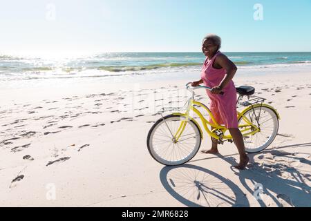Carefree african american senior woman with bicycle standing on sandy beach against clear sky Stock Photo