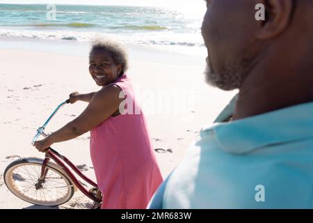 Happy african american senior woman with bicycle walking at beach and talking with man on sunny day Stock Photo