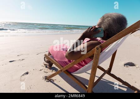 African american senior woman talking over mobile phone while relaxing on deckchair at beach Stock Photo
