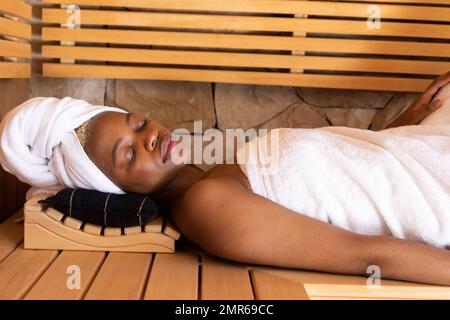 Happy african american woman wearing towel and sitting in sauna