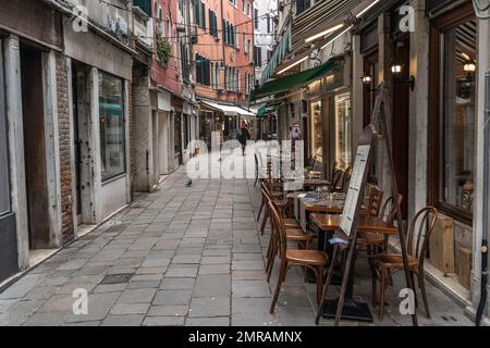 Empty tables of outdoor cafe at the empty narrow street in the old town of Venice, Italy at winter with one person walking in a distance. Vintage tabl Stock Photo