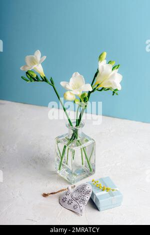 Gift box, vintage toy heart and freesia white flowers in glass vase on white textured table against blue wall. Valentines day, holiday concept. Closeu Stock Photo