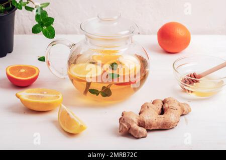Glass teapot with orange, lemon, ginger and mint tea with ingredients on white wooden table. Stock Photo