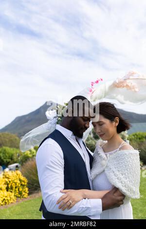 Vertical of happy, diverse bride and groom embracing with eyes closed at outdoor wedding, copy space Stock Photo