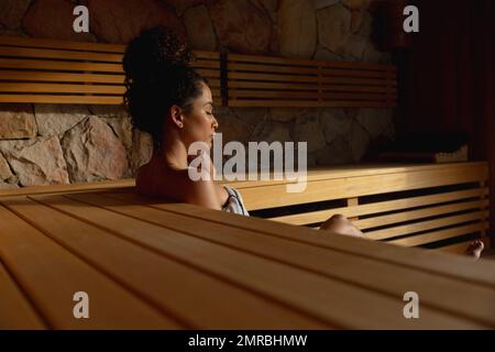 Tranquil biracial woman wearing towel relaxing with eyes closed in sauna, copy space Stock Photo