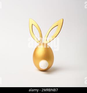 Golden Easter egg with bunny ears and tail on white background. Minimal style, side view. Happy Easter concept. Stock Photo
