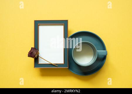 Empty navy photo frame and dry rose flower, cup of milk on yellow background. top view, copy space Stock Photo