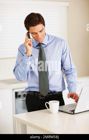 Young executive using cellphone and laptop while at the kitchen. Young business man using cellphone and laptop while at the kitchen. Stock Photo