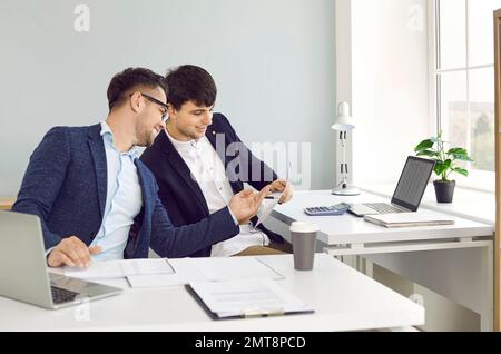 Two male office workers are reviewing documents together while sitting at adjacent desks in office. Stock Photo