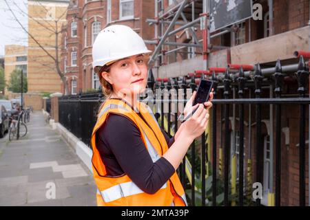 Chartered civil engineer lady showing you something on a smart phone with electronic pen, construction site, hard hat and orange high visibility vest, Stock Photo