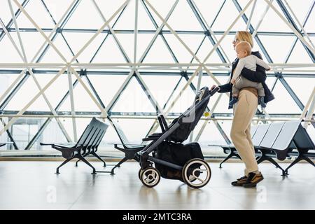 Mother carying his infant baby boy child, pushing stroller at airport departure terminal moving to boarding gates to board an airplane. Family travel with baby concept Stock Photo