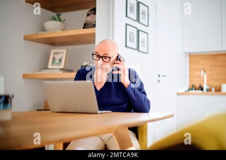 Wondered middle aged man making a call and using laptop while working from home. Confident male sitting in the dining room and having a call. Home off Stock Photo