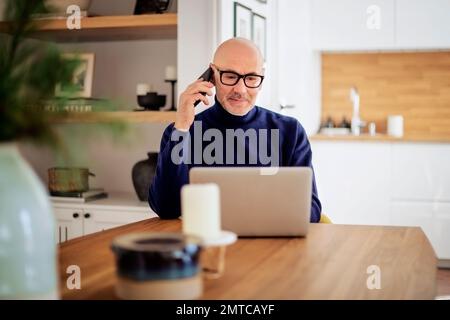 Confident middle aged man making a call and using laptop while working from home. Confident male sitting in the dining room and having a call. Home of Stock Photo