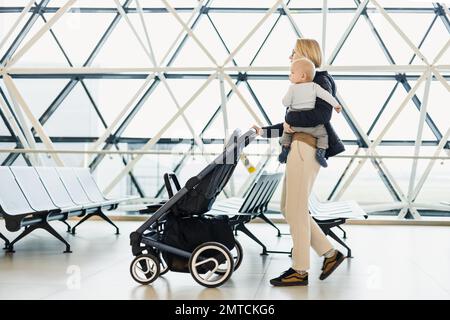 Mother carying his infant baby boy child, pushing stroller at airport departure terminal moving to boarding gates to board an airplane. Family travel with baby concept Stock Photo