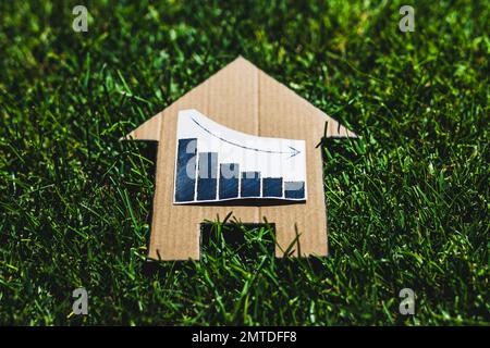 interest rates going down, graph showing stats decreasing over house icon made of cardboard on green lawn Stock Photo