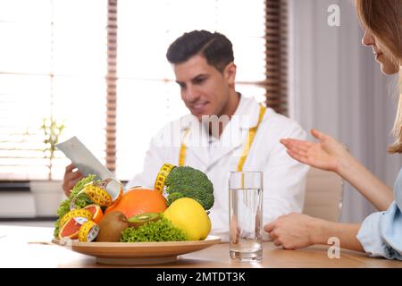 Nutritionist consulting patient at table in clinic, focus on plate with fruits, vegetables and measuring tape Stock Photo
