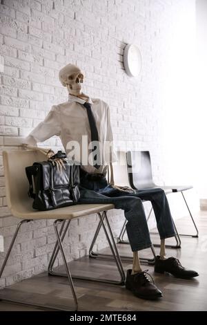 Human skeleton in office wear sitting on chair near brick wall indoors Stock Photo