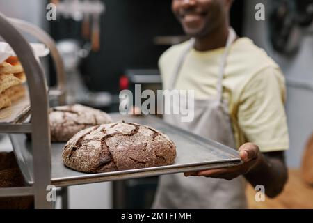 Closeup image of smiling baker putting tray with freshly baked bread in bakers rack Stock Photo