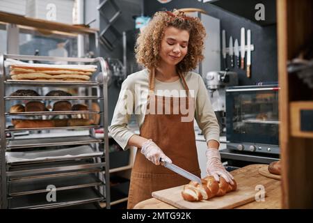 Smiling baker wearing gloves when cutting bread for customers Stock Photo