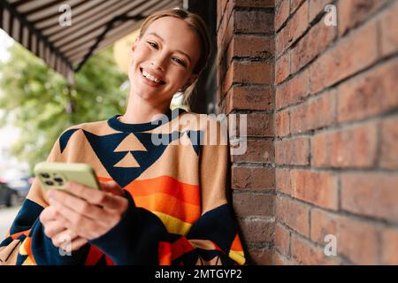Pretty joyful woman in colorful sweater using smartphone while standing near brick wall at city street Stock Photo