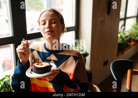 Young woman in colorful sweater eating delicious dessert while sitting at table in cafe Stock Photo
