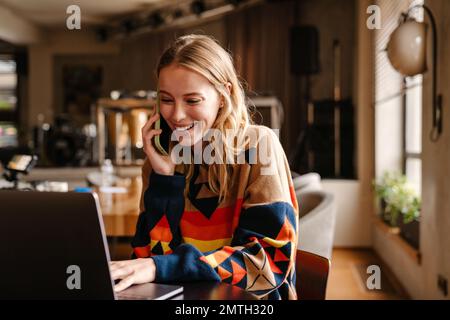 Excited cheerful young woman wearing colorful sweater talking on cellphone while using laptop sitting at table in cafe Stock Photo