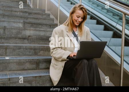 Young beautiful happy smiling business lady working with laptop, while sitting on the stairs outdoors Stock Photo