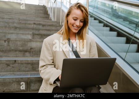 Young beautiful happy smiling business lady working with laptop, while sitting on the stairs outdoors Stock Photo