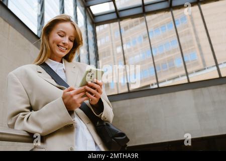 Young beautiful smiling happy woman holding and using her phone, while standing indoors leaning on the railing Stock Photo