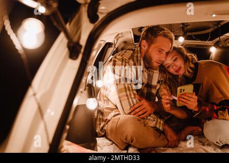 Happy young white couple using cellphone and lying together in car outdoors Stock Photo