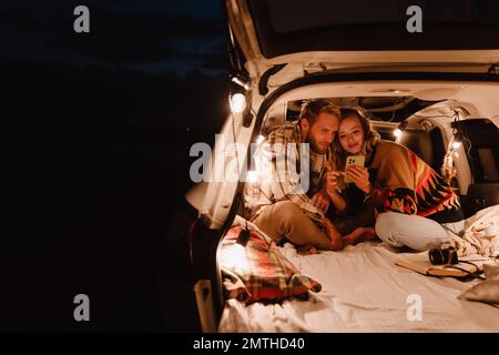 Happy young white couple using cellphone and lying together in car outdoors Stock Photo