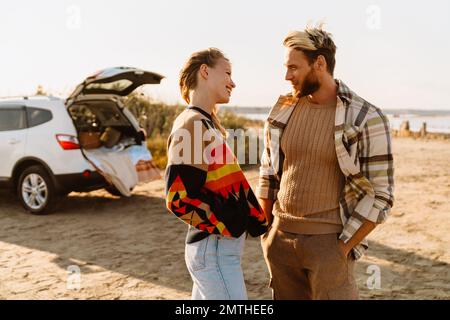Happy young white couple smiling and looking at each other while walking by seashore Stock Photo