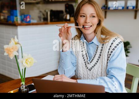 Young beautiful long-haired smiling woman in shirt and vest biting pencil and looking aside while doing her homework with laptop in cozy cafe Stock Photo