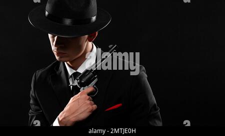 Stylish portrait of gangster from 1940s with a gun. Man in a black suit and hat with a gun over dark background. Stock Photo