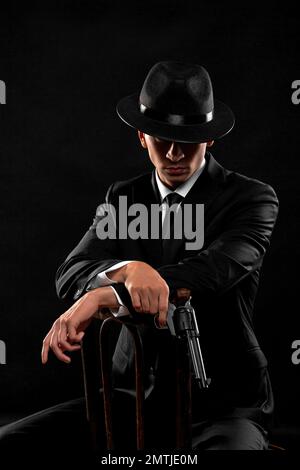 Vertical portrait of gangster from 1940s with a gun. Man in a black suit and hat sitting with a gun over black background. Stock Photo