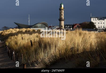 01 February 2023, Mecklenburg-Western Pomerania, Warnemünde: The beach grass on the dunes shines brightly in the sun, while dark storm clouds pass behind the lighthouse and the Teepott. Photo: Bernd Wüstneck/dpa Stock Photo