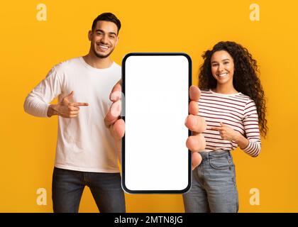 Nice Offer. Smiling Middle Eastern Couple Pointing At Big Blank Smartphone Stock Photo