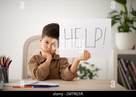 Boring tired sad small european boy sits at table shows paper with inscription help in school room interior Stock Photo