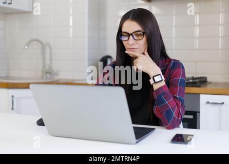 Pensive young woman in glasses thinking about problem solution in front of laptop computer. Freelancer female works at home Stock Photo