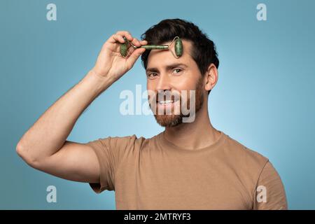 Portrait of handsome man using face roller for smooth and firm anti aging skin treatment, blue background Stock Photo